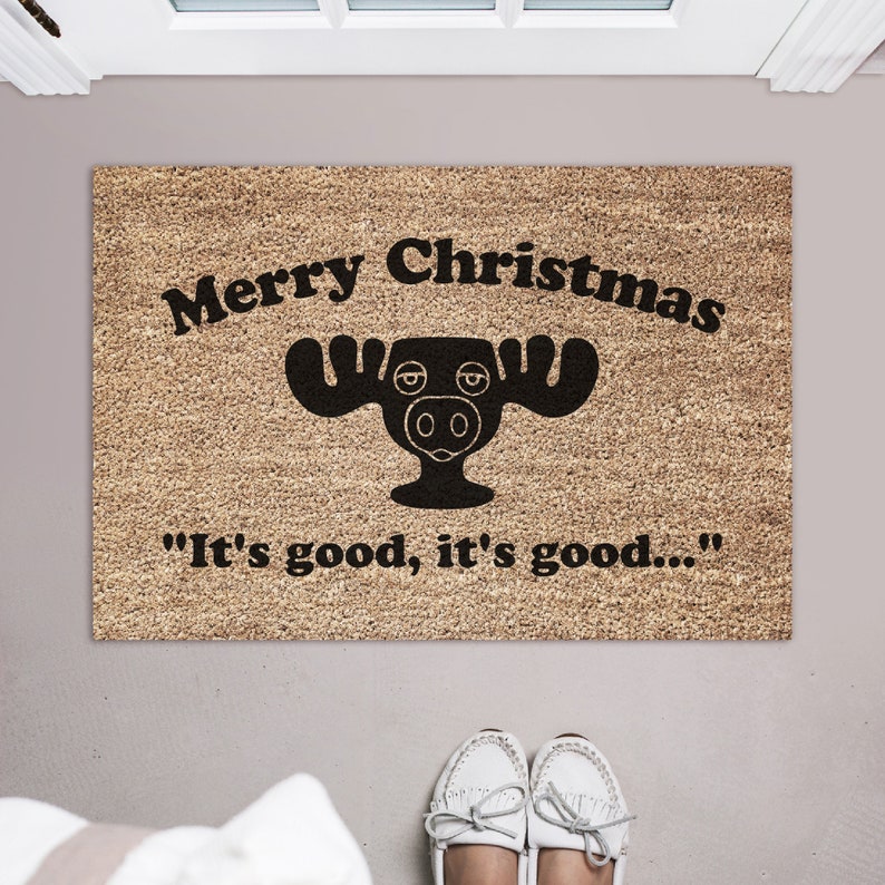 May include: A brown doormat with the text "Merry Christmas" and a black cartoon moose with the text "It's good, it's good..." underneath.