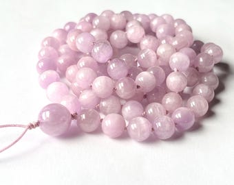 May include: A strand of light purple gemstone beads, likely amethyst, arranged in a circular pattern. The beads are round and smooth, with a soft, translucent appearance. A larger bead is at the end, with a pink string attached.