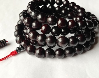 May include: A black wooden mala bead necklace with a red string tassel. The beads are round and smooth, and the necklace is approximately 108 beads long.
