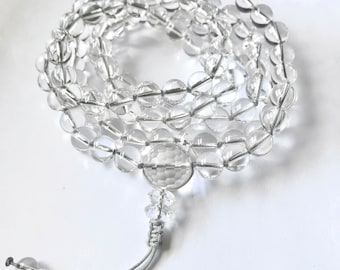 May include: A clear quartz crystal mala necklace with a large, faceted crystal bead at the center. The necklace is made of 108 beads and has a tassel at the end.