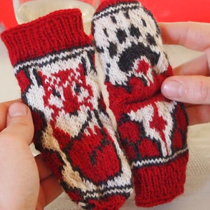 Op de afbeelding: Een paar rode, witte en grijze gebreide vingerloze handschoenen met een geometrisch patroon. De handschoenen hebben een pootprintontwerp op de ene handschoen en een gestileerd dierenontwerp op de andere.