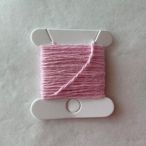 May include: A white plastic bobbin wrapped with light pink yarn. The bobbin features a rectangular center section with rounded ends and a small circular cutout at the bottom. The yarn is wound horizontally across the center of the bobbin.