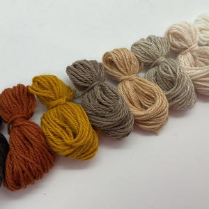 Puede incluir: Una colección de madejas de hilo en una variedad de tonos cálidos y terrosos. Los colores van desde el negro intenso y óxido hasta el amarillo mostaza, beige y crema. Cada madeja está cuidadosamente empaquetada y atada, ideal para proyectos de manualidades.