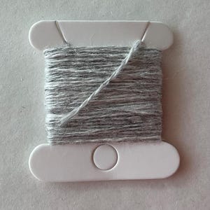 May include: A white plastic bobbin wrapped with light gray, textured yarn. The bobbin features a flat, rectangular shape with rounded ends and a circular cutout in the center of the bottom section, with the yarn wound horizontally across the middle.