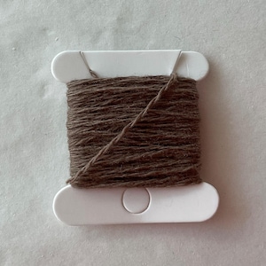 May include: A white plastic spool wrapped with brown thread. The thread is wound horizontally around the center of the spool, with a single diagonal strand crossing the middle section. The spool features rounded ends and a circular cutout in the center.