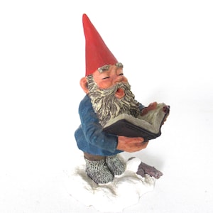 May include: A figurine of a garden gnome with a red pointed hat and blue jacket, singing from a book. The gnome has a long white beard and is standing on a white cloud-like base. The book has dark brown covers.