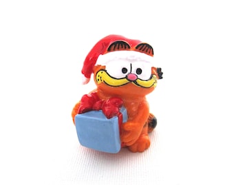 May include: A small orange and white figurine of Garfield the cat wearing a red Santa hat and holding a blue gift box with a red bow.