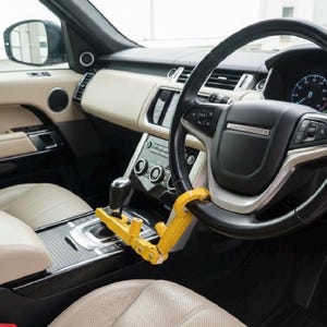 May include: A bright yellow steering wheel and gear shift lock device installed inside a vehicle. The security tool connects the black leather steering wheel to the center console gear shifter, featuring a metal frame and a locking mechanism.