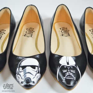 May include: Three pairs of black ballet flats with pointed toes. The flats are painted with Star Wars characters: Darth Vader, a Stormtrooper, and a TIE Fighter. The flats have a white interior and the word "LUW" printed on the inside.