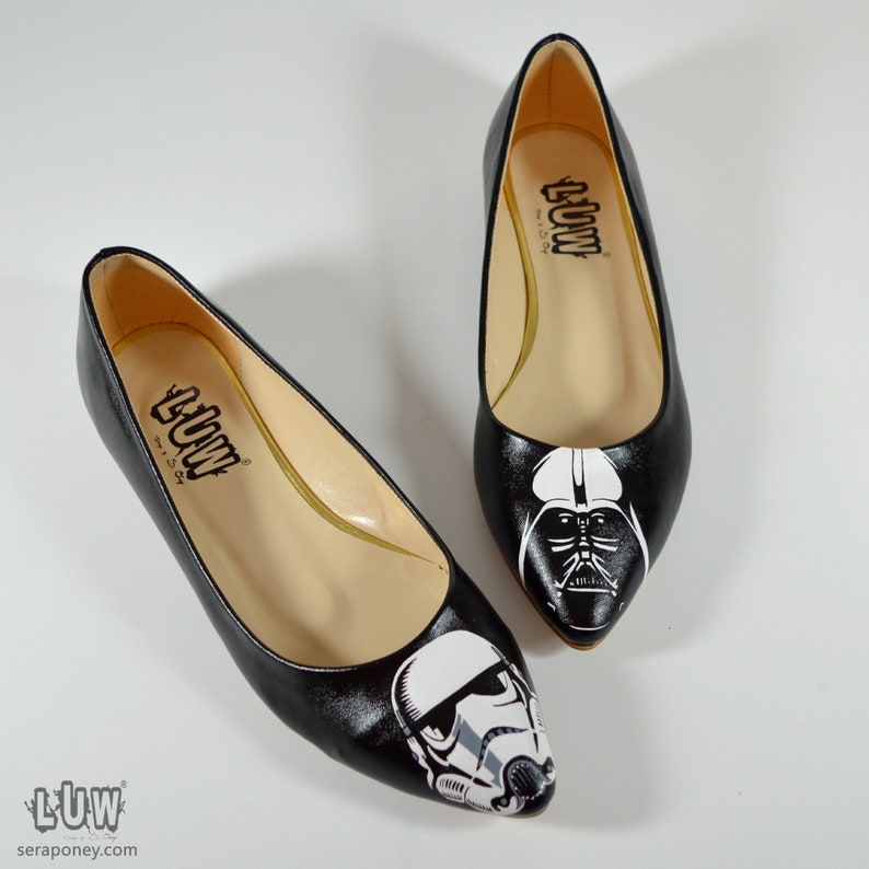 May include: A pair of black flats with pointed toes. One flat features a white and black illustration of Darth Vader, while the other flat features a white and black illustration of a Stormtrooper. The flats have a black leather upper and a beige insole. The insole has the word "LUW" printed on it.