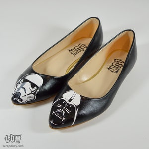 May include: A pair of black ballet flats with a pointed toe. One flat features a white and black illustration of Darth Vader, while the other flat features a white and black illustration of a Stormtrooper. The flats have the word "LUW" printed on the insole.