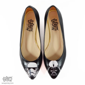 May include: A pair of black ballet flats with a pointed toe. One flat features a white and black illustration of a Stormtrooper helmet, while the other flat features a white and black illustration of Darth Vader's helmet. The flats have a gold trim around the inside of the shoe and the text "LUW" is printed on the inside of each shoe.
