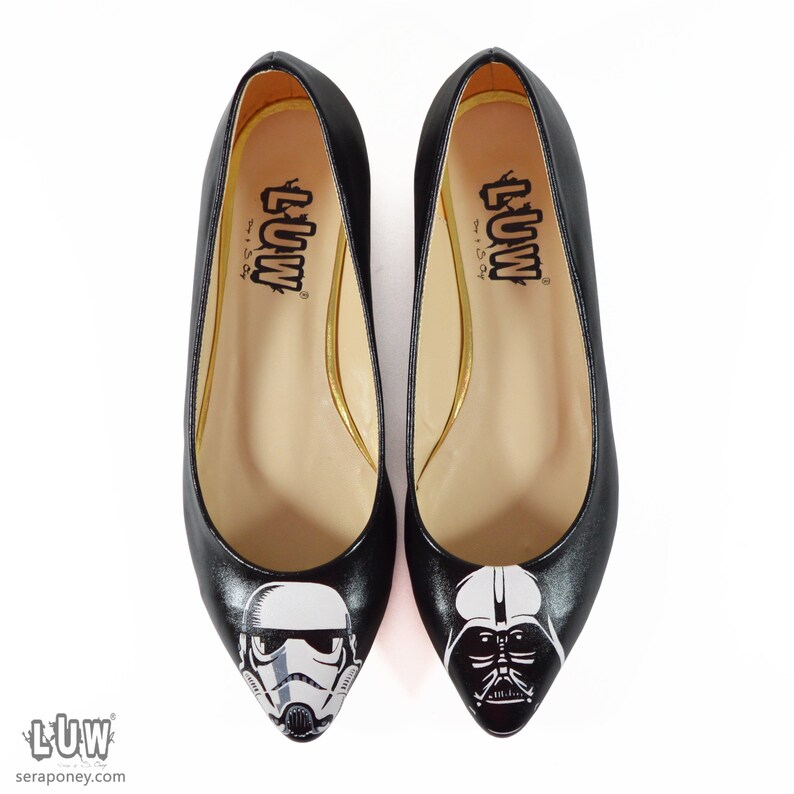 May include: A pair of black ballet flats with a pointed toe. One flat features a white and black illustration of a Stormtrooper helmet, while the other flat features a white and black illustration of Darth Vader's helmet. The flats have a gold trim around the inside of the shoe and the text "LUW" is printed on the inside of each shoe.