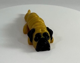 May include: A small, golden brown, 3D printed figurine of a dog. The dog is lying down with its head resting on the ground. The dog has black eyes and a black nose.