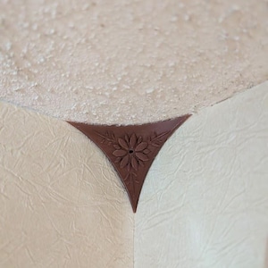 Könnte beinhalten: Braune, dreieckige Eckleiste mit floralem Design. Das dekorative Element ist dort angebracht, wo sich zwei Wände treffen, mit einer strukturierten Decke im Hintergrund. Die Leiste hat eine dunkelrotbraune Farbe.