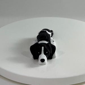 Puede incluir: Una figurita de perro acostado impresa en 3D en blanco y negro. El perro tiene la nariz negra y los ojos negros.