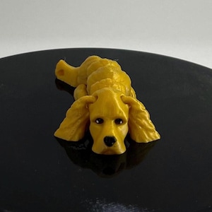 Puede incluir: Una pequeña figurita de un perro acostado, impresa en 3D, de color dorado. El perro tiene orejas caídas y una nariz negra.