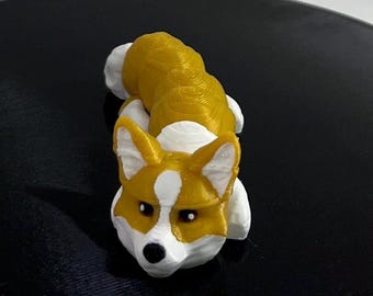 May include: A 3D printed figurine of a corgi dog. The dog is lying down with its head raised and looking at the camera. The figurine is made of a light brown plastic with white markings.