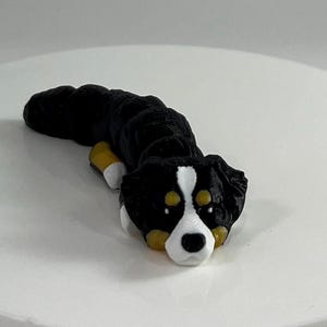 Peut inclure: Une figurine de chien de montagne bernois noir et blanc avec des marques brunes autour des yeux. La figurine est enroulée dans une position de sommeil.