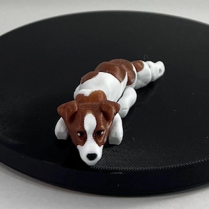 Può includere: Una piccola statuina di un cane sdraiato, stampata in 3D, di colore marrone e bianco, con la testa appoggiata sulle zampe.