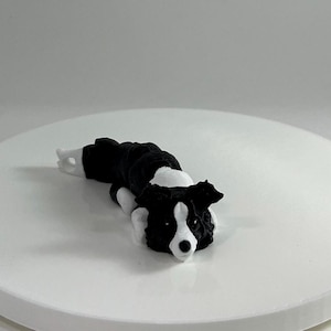 May include: A black and white border collie figurine lying on its side with its head resting on its paws. The figurine is made of a smooth, matte material.
