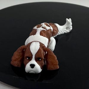 Op de afbeelding: Een klein, bruin en wit beeldje van een liggende Cavalier King Charles Spaniel. Het beeldje is gemaakt van plastic en heeft een realistisch uiterlijk.