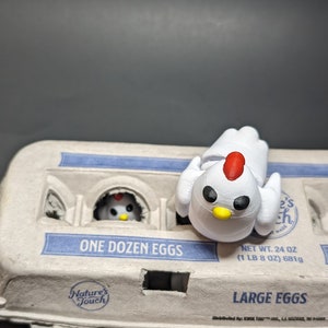 May include: A white ceramic chicken figurine with black eyes and a red comb sits on top of a cardboard egg carton. The carton is labelled "ONE DOZEN EGGS" and "LARGE EGGS".