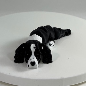 May include: A black and white 3D printed figurine of a dog lying down. The dog has floppy ears and a white patch on its chest.