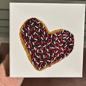 Può includere: Un'opera d'arte su tela a forma di cuore con un design di perline che ricorda una ciambella glassata al cioccolato con zuccherini. La ciambella ha un centro marrone, un bordo giallo-oro ed è decorata con zuccherini rossi, rosa e bianchi. La tela è quadrata e bianca.