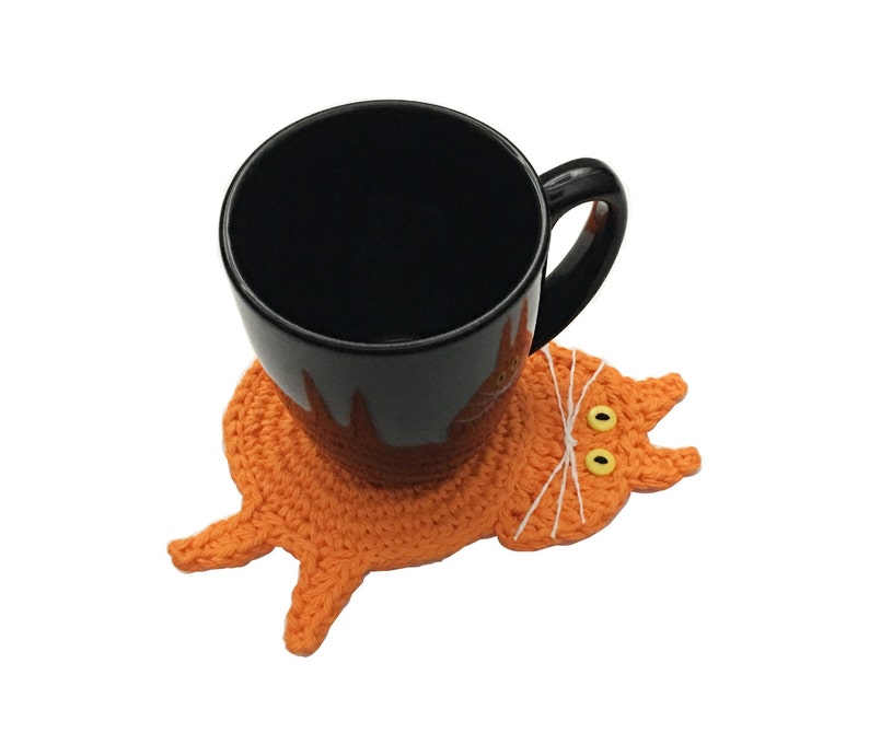 Crochet cat butt coaster on display with a coffee cup on top. Coaster measures at approximately 3.5-4"W across the body (excluding head, tail and legs). Coaster width is great for most coffee cups, drink glasses and water bottles.