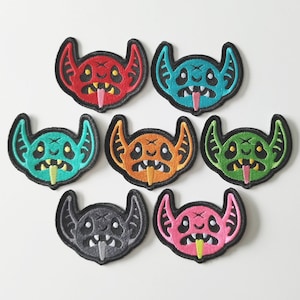 May include: Eight embroidered iron-on patches featuring a cartoon monster with a toothy grin and a long tongue. The patches are in various colors, including red, orange, green, blue, pink, and gray.