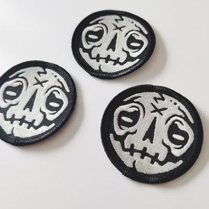 May include: Three black and white embroidered iron-on patches with a cartoon skull design. The skull has a cross on its forehead and a stitched-up mouth.