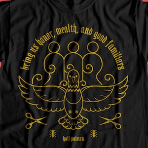 May include: Black t-shirt with a gold graphic design. The design includes a bird with outstretched wings, three figures, scissors, and the text "bring us honor, wealth, and good familiars." The text "hail paimon" is also included.