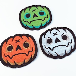 Puede incluir: Tres parches bordados de Halloween con forma de calabazas. Uno es naranja, otro verde y otro blanco. Cada uno tiene un contorno negro y una cara cosida con una cruz sobre los ojos. Los parches están sobre un fondo blanco.