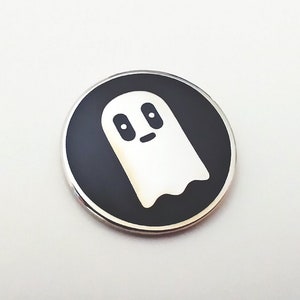 May include: A black and white enamel pin featuring a cartoon ghost with a smiling face. The ghost is white with black eyes and a mouth. The pin is circular and has a silver metal border.