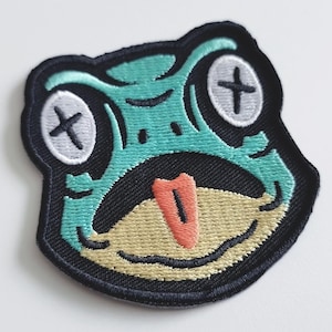 Op de afbeelding: Een geborduurde patch met een groen kikkergezicht met zwarte stiksels en witte ogen met zwarte X's. De kikker heeft een roze tong die uitsteekt.