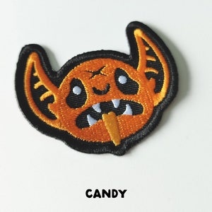 Pu&ograve; includere: Toppa ricamata in ferro di colore arancione e nero con un mostro dei cartoni animati con zanne e un bastoncino di caramelle in bocca. Il testo "CANDY" &egrave; sotto la toppa.