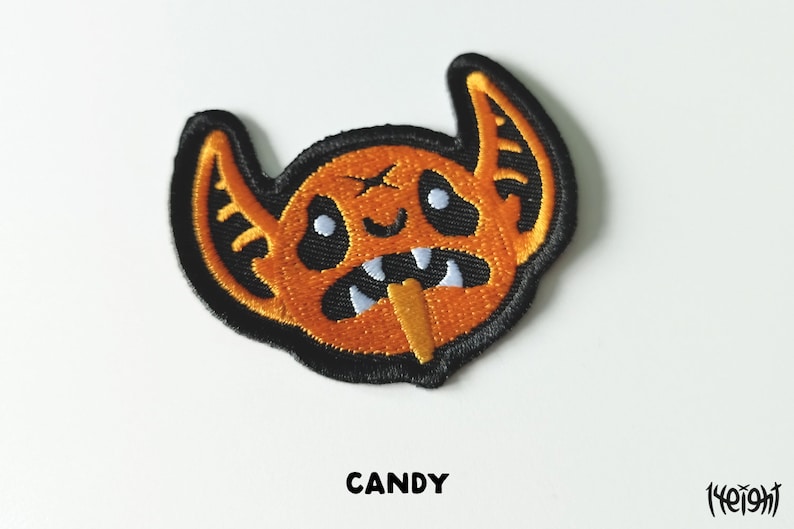Pu&ograve; includere: Toppa ricamata in ferro di colore arancione e nero con un mostro dei cartoni animati con zanne e un bastoncino di caramelle in bocca. Il testo "CANDY" &egrave; sotto la toppa.