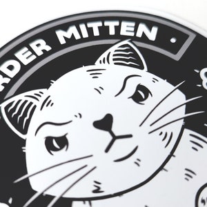 Pu&ograve; includere: Adesivo rotondo in bianco e nero con un'illustrazione di un gatto e il testo "MURDER MITTEN." Il gatto ha un'espressione maliziosa e guarda direttamente lo spettatore. L'adesivo ha un bordo grigio.