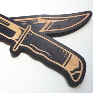 May include: Two black wooden knives with a light brown wood grain pattern. The knives are crossed over each other.