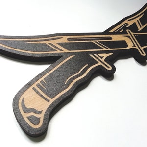 May include: Two black wooden knives with a brown wood grain design are crossed over each other. The knives are outlined in black and have a detailed design.