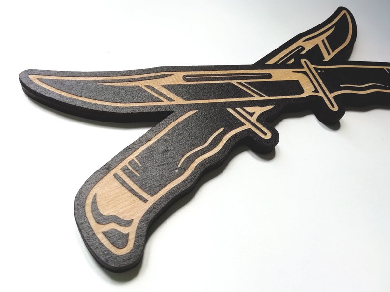 May include: Two black wooden knives with a brown wood grain design are crossed over each other. The knives are outlined in black and have a detailed design.