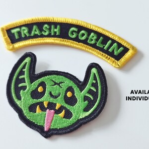 Pu&ograve; includere: Due toppe ricamate, una con una creatura verde simile a un goblin con una lingua rosa e l'altra con uno stendardo giallo che recita "Trash Goblin".
