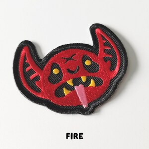 Pu&ograve; includere: Toppa ricamata rossa con un viso di diavolo dei cartoni animati con occhi e denti gialli e una lingua rosa che sporge. La toppa ha cuciture nere intorno ai bordi. La parola "FIRE" &egrave; stampata sotto la toppa.