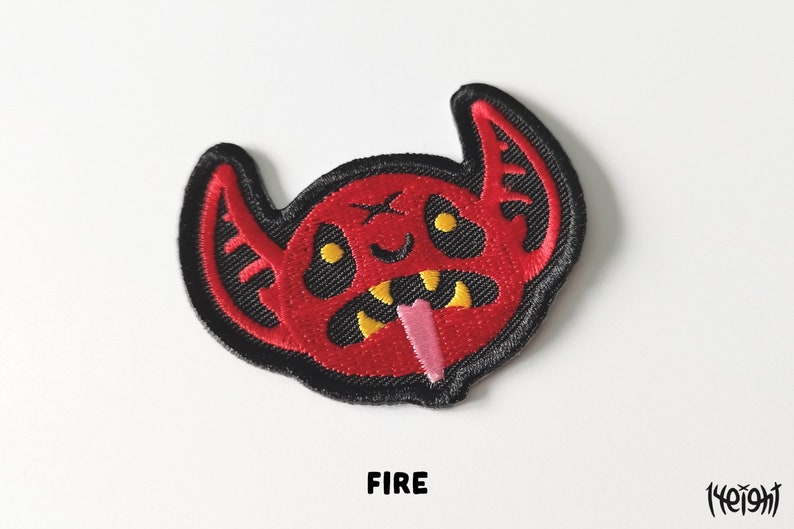 Pu&ograve; includere: Toppa ricamata rossa con un viso di diavolo dei cartoni animati con occhi e denti gialli e una lingua rosa che sporge. La toppa ha cuciture nere intorno ai bordi. La parola "FIRE" &egrave; stampata sotto la toppa.