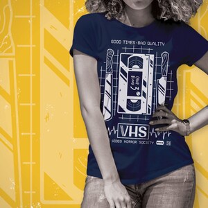 May include: A navy blue t-shirt with a white graphic of a VHS tape and knives. The text on the tape reads "Camp 3: Bloody", and the text below the graphic reads "VHS Video Horror Society".