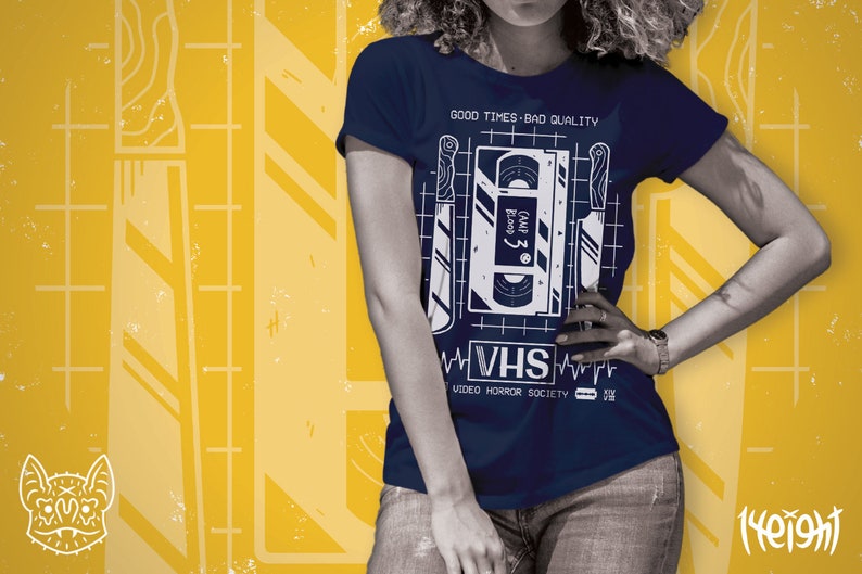 May include: A navy blue t-shirt with a white graphic of a VHS tape and knives. The text on the tape reads "Camp 3: Bloody", and the text below the graphic reads "VHS Video Horror Society".