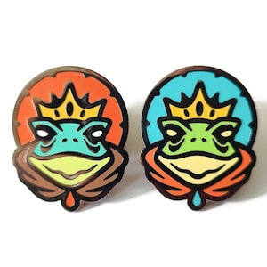 May include: Two circular enamel pins featuring a stylised frog wearing a yellow crown. The left pin has an orange background, while the right pin has a light blue background. Both frogs are green with dark outlines and a small teardrop shape below.