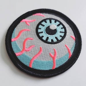 Peut inclure: Un patch brodé rond, bordé de noir, représentant un œil de dessin animé avec un iris bleu, une pupille blanche et des veines roses.