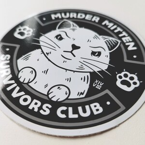 Pu&ograve; includere: Adesivo bianco e nero con un gatto dei cartoni animati con un'impronta di zampa e il testo "Murder Mitten Survivors Club".
